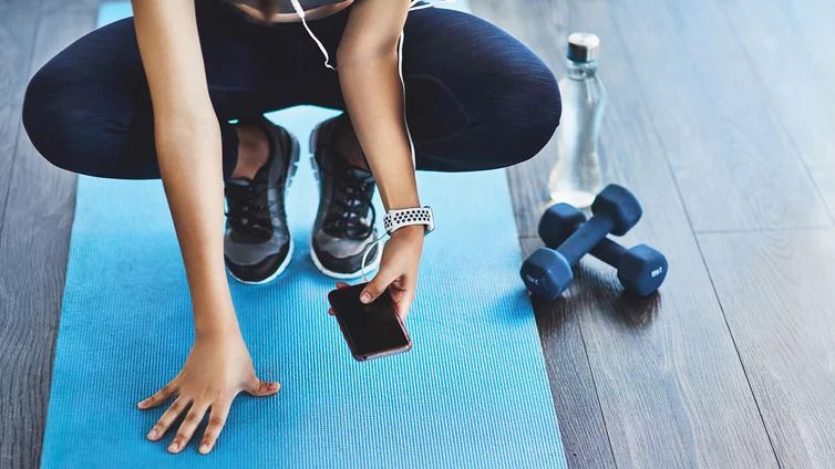 Person in Sportkleidung sitzt auf einer blauen Yogamatte und hält ein Smartphone in der Hand, neben ihr liegen zwei Hanteln und eine Wasserflasche auf dem Holzboden