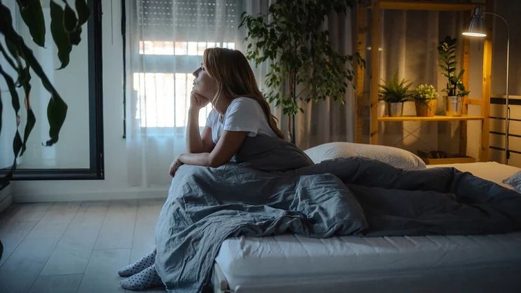 Person sitzt auf einem Bett, hält ein Kissen und blickt aus einem Fenster in einem gemütlich eingerichteten Zimmer mit Pflanzen und Holzregal